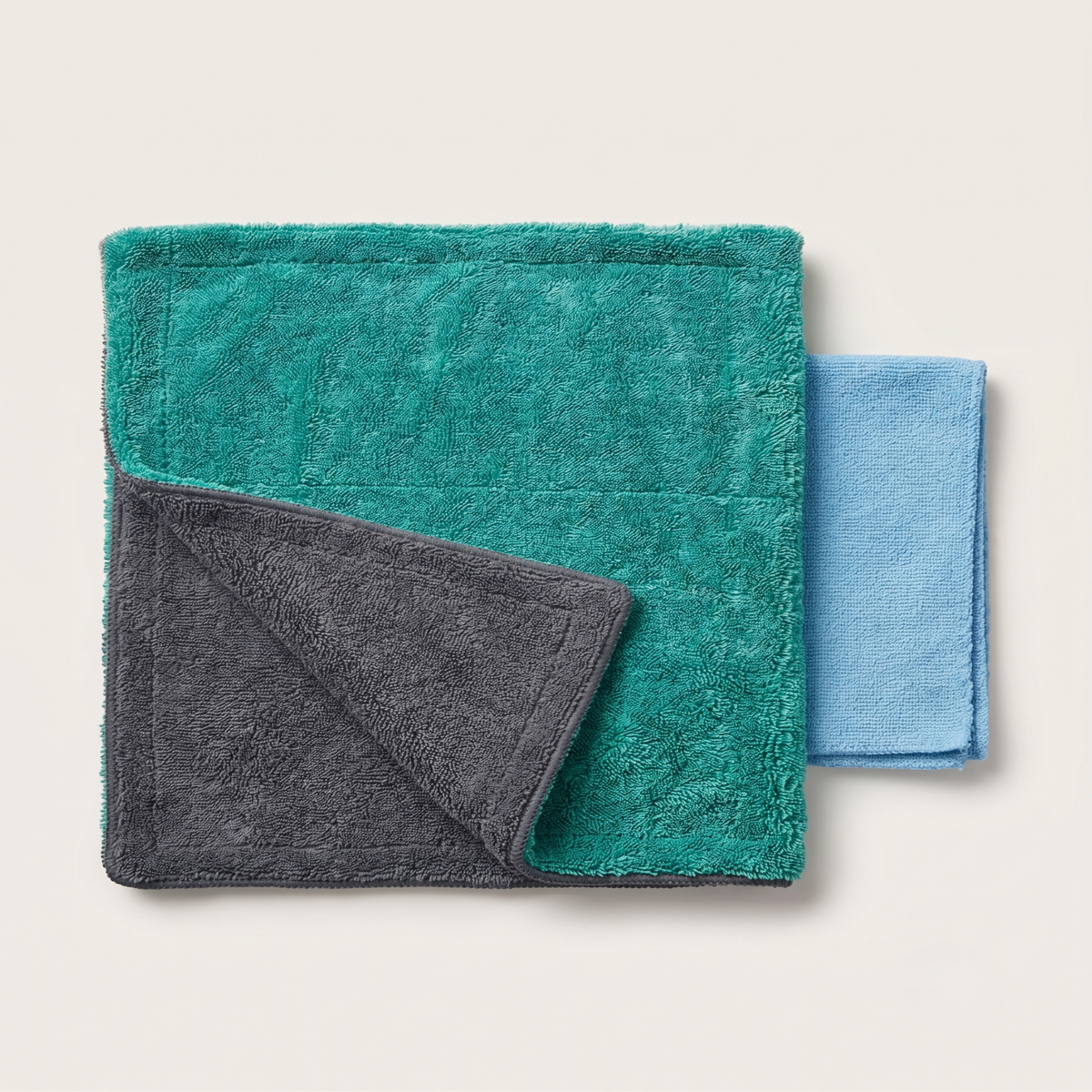 Two folded plush towels or blankets on a light warm gray background: a larger teal-green towel with a dark charcoal underside, and a smaller light blue towel partially visible behind it.